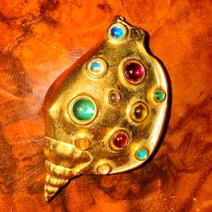 May include: A gold-toned seashell-shaped brooch with colorful gemstones. The brooch features a variety of round gemstones in shades of blue, red, green, and yellow. The brooch is set against a warm, reddish-brown background.
