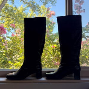 May include: A pair of black knee-high boots with a smooth finish, rounded toes, and a block heel. The boots feature a tall shaft that reaches below the knee, a structured silhouette, and a clean design without visible zippers or decorative hardware.
