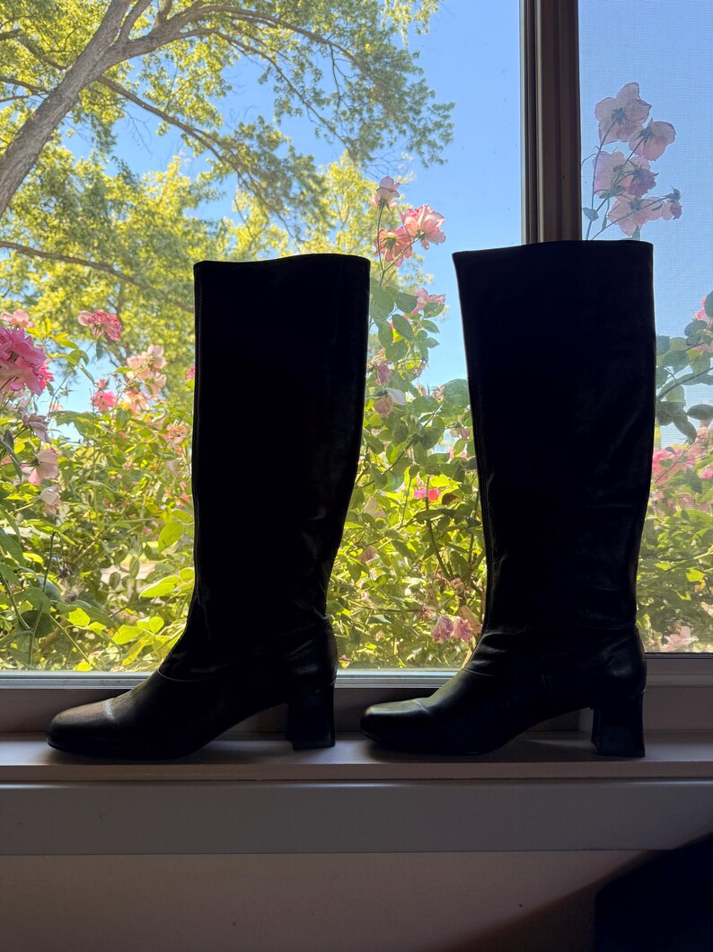 May include: A pair of black knee-high boots with a smooth finish, rounded toes, and a block heel. The boots feature a tall shaft that reaches below the knee, a structured silhouette, and a clean design without visible zippers or decorative hardware.