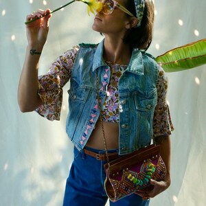 Puede incluir: Una mujer lleva un chaleco vaquero con bordados rosa y azul, una blusa de estampado floral, unos vaqueros azules con adornos en forma de coraz&oacute;n y un bolso marr&oacute;n y multicolor.