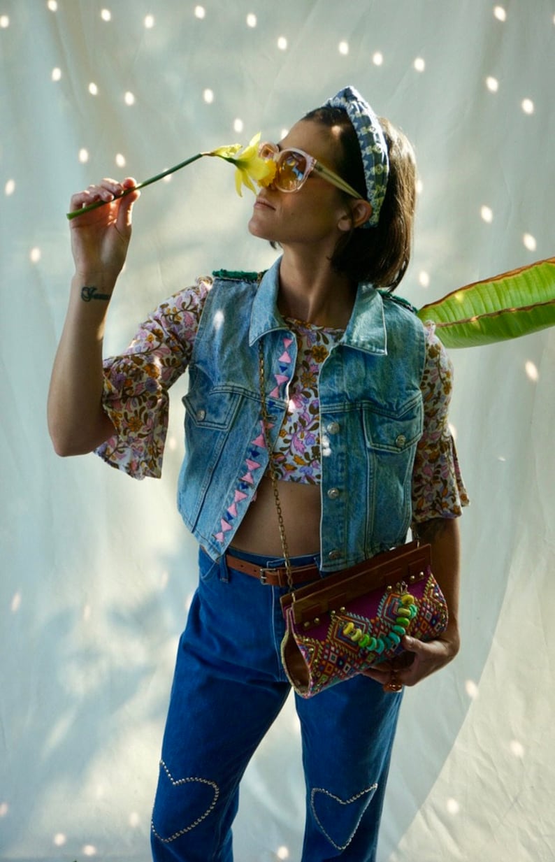Puede incluir: Una mujer lleva un chaleco vaquero con bordados rosa y azul, una blusa de estampado floral, unos vaqueros azules con adornos en forma de coraz&oacute;n y un bolso marr&oacute;n y multicolor.