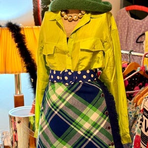 May include: A green felt cloche hat, a gold-toned beaded necklace, a lime green button-down shirt, a blue belt with white polka dots, and a blue, green, and white plaid skirt with a fringed edge and four visible buttons on the side.