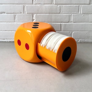 May include: An orange dice-shaped desk organiser with red and black accents. The organiser dispenses white paper from a cylindrical opening. The background is a white brick wall.