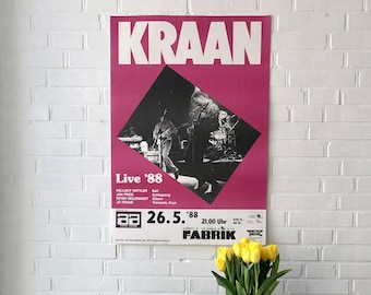 Op de afbeelding: Een witte platenspeler met een zwarte draaitafel staat op een wit metalen rek met een platencollectie. Achter de platenspeler hangt een poster voor een KRAAN-concert op 26 mei 1988 in de FABRIK in Berlijn, Duitsland. Een vaas met gele bloemen staat op het rek.