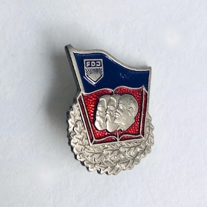 May include: A vintage pin featuring a blue flag with the letters "FDJ" and a shield emblem. Below the flag is a red section with two faces and an open book. The pin is surrounded by a silver wreath.