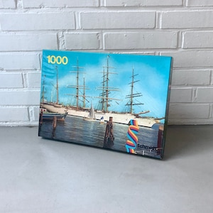 Può includere: Una scatola di puzzle da 1000 pezzi con una grande nave a vela bianca con una vela a strisce blu e bianche in un porto. La scatola è blu con un nastro arcobaleno e il testo "Schmid" nell'angolo in basso a destra.