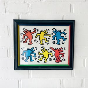 May include: Framed artwork featuring Keith Haring-style figures in red, yellow, and blue on a white background. The figures are dancing and framed in a black frame with a green and red border. The artwork is displayed against a white brick wall.