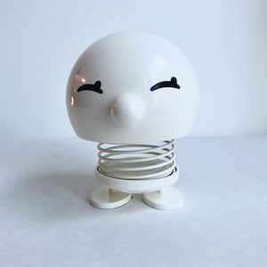 May include: A white ceramic spring-loaded toy with a smiling face and black eyes. The toy has a spring coil and two small feet.