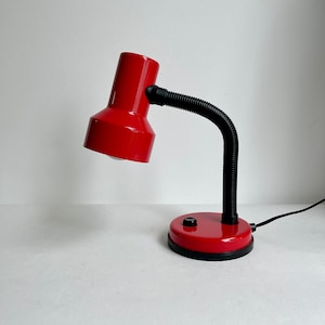 May include: A red desk lamp with a flexible black arm and a round base. The lamp has a white light bulb and a black on/off switch.