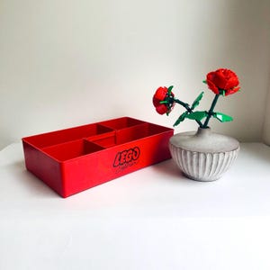 Könnte beinhalten: Eine rote LEGO System Aufbewahrungsbox mit Innenteilern. Das LEGO-Logo ist schwarz auf der Seite aufgedruckt. Daneben steht eine graue Vase mit zwei roten LEGO-Rosen.