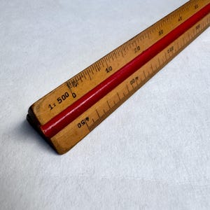 May include: A triangular wooden scale ruler featuring a central red groove and black printed markings. The ruler displays various numerical scales, including 1:500, with clear graduation lines along the edges for precise technical measurements.