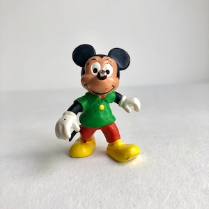 Puede incluir: Una figurita de plástico vintage de Mickey Mouse. Lleva una camisa verde con un botón amarillo, pantalones rojos y zapatos amarillos. Tiene orejas negras y una nariz negra.