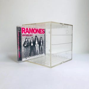 May include: A transparent acrylic CD storage cube with a CD case featuring the band Ramones. The CD case is partially inserted into the cube, displaying the album cover with the band members in front of a brick wall.