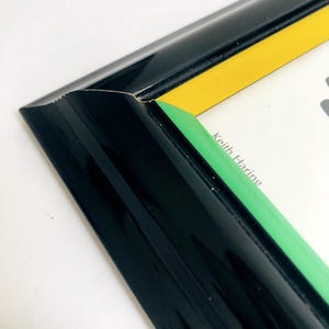 May include: Close-up of a black picture frame with yellow and green accents. The frame has a glossy finish and a clean, modern design. The text "Keith Haring" is visible on the artwork within the frame.