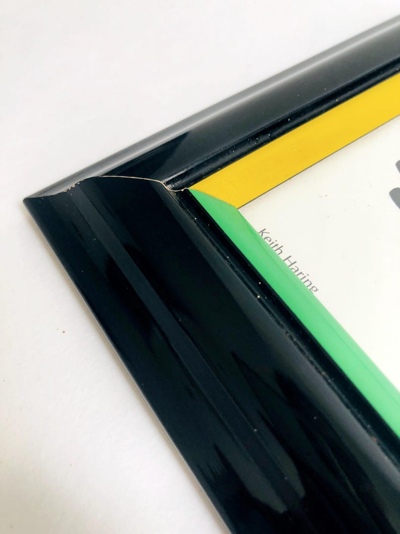 May include: Close-up of a black picture frame with yellow and green accents. The frame has a glossy finish and a clean, modern design. The text "Keith Haring" is visible on the artwork within the frame.
