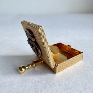 Könnte beinhalten: Kleine, offene, goldfarbene Metallbox mit einem Netzmuster. Die Box hat einen Klappdeckel mit dekorativem Relief und einen kleinen, runden, goldfarbenen Gegenstand im Inneren. Ein goldfarbener Griff erstreckt sich von der Seite und endet in einer runden Kugel.
