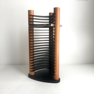 May include: A vertical media storage rack featuring three light brown wooden cylindrical pillars connected by a black triangular base. The rack includes multiple black curved horizontal shelves designed to hold compact discs, with one CD case inserted.