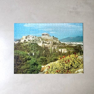 K&ouml;nnte beinhalten: Ein Puzzle, das eine malerische Aussicht auf die Akropolis in Athen, Griechenland, zeigt. Das Puzzle zeigt ein detailliertes Bild des antiken griechischen Wahrzeichen, einschlie&szlig;lich des Parthenon, des Erechtheion und der umliegenden H&uuml;gel. Die Puzzleteile sind in einem Raster angeordnet, und das Bild ist von einem wei&szlig;en Rand umgeben.