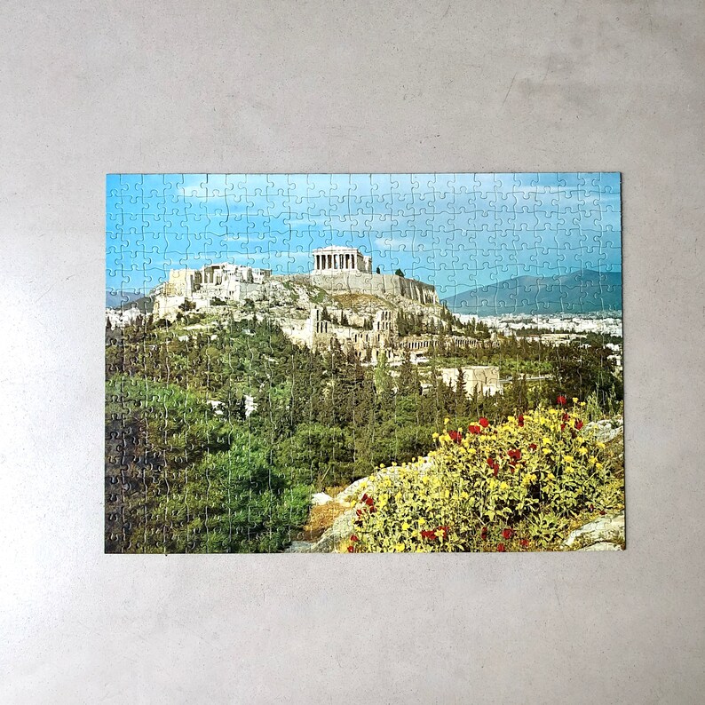 K&ouml;nnte beinhalten: Ein Puzzle, das eine malerische Aussicht auf die Akropolis in Athen, Griechenland, zeigt. Das Puzzle zeigt ein detailliertes Bild des antiken griechischen Wahrzeichen, einschlie&szlig;lich des Parthenon, des Erechtheion und der umliegenden H&uuml;gel. Die Puzzleteile sind in einem Raster angeordnet, und das Bild ist von einem wei&szlig;en Rand umgeben.