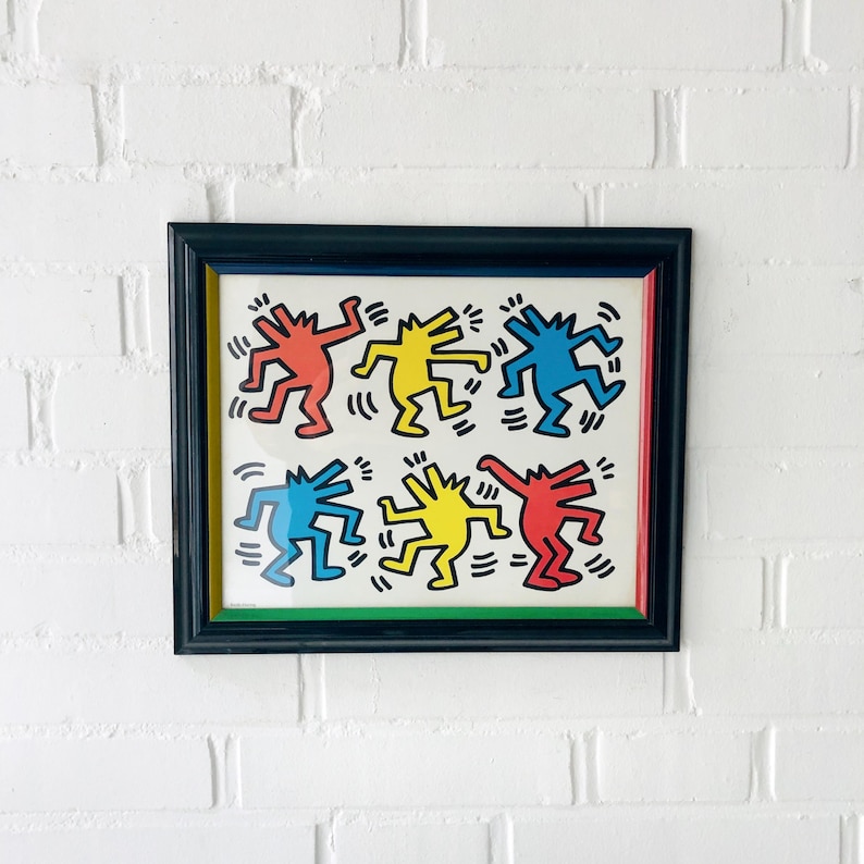 May include: Framed artwork featuring Keith Haring-style figures in red, yellow, and blue on a white background. The figures are dancing and framed in a black frame with a green and red border. The artwork is displayed against a white brick wall.