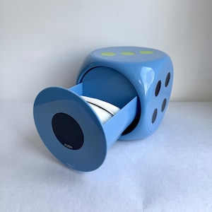 May include: A light blue, die-shaped container with a pull-out drawer. The drawer is open, revealing white items inside. The die has black dots on the sides and green dots on top. The drawer has a black circle with the word "PUSH".