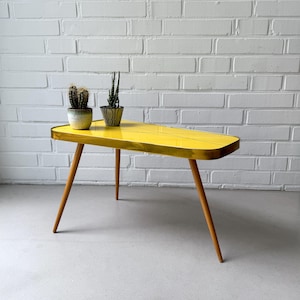 Op de afbeelding: Een gele salontafel in retrostijl met een gouden rand en drie houten poten. De tafel heeft een kenmerkende, langwerpige vorm en is versierd met zwarte lijnen. Twee kleine cactussen in potten staan op het oppervlak.