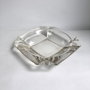 Könnte beinhalten: Ein Aschenbecher aus klarem Glas mit quadratischer Form und leicht erhöhtem Rand. Der Aschenbecher hat ein schlichtes Design ohne Verzierungen.