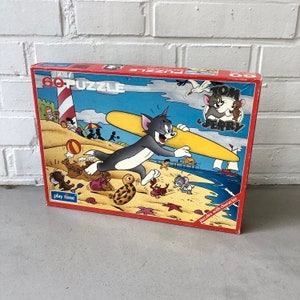 Könnte beinhalten: Ein 60-teiliges Tom und Jerry Puzzle mit Tom auf einem Surfbrett am Strand. Die Schachtel ist rot und zeigt ein Cartoon-Bild von Tom und Jerry.
