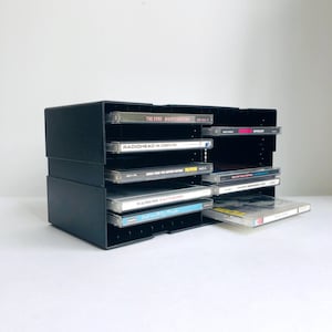 May include: A black CD storage unit filled with CDs. The unit is made of a sturdy material and has multiple compartments for organising CDs. The CDs are visible, with their album titles and artwork displayed. The unit is designed to hold a collection of CDs.