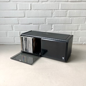May include: A black CD storage case with a sliding door and a hinged front panel. The case is rectangular and holds several CDs. The open door reveals a collection of CD cases. The case is on a white surface.