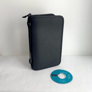 May include: A black CD case with a zip closure stands upright beside a compact disc. The CD is teal with white text and the band name "Oasis". The case is rectangular and has a textured surface.