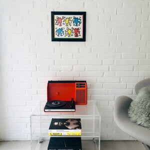 May include: A framed artwork with colorful dancing figures hangs on a white brick wall. Below, a clear acrylic rolling cart holds a vintage orange record player and a stack of books. A gray armchair with a fluffy pillow sits nearby.