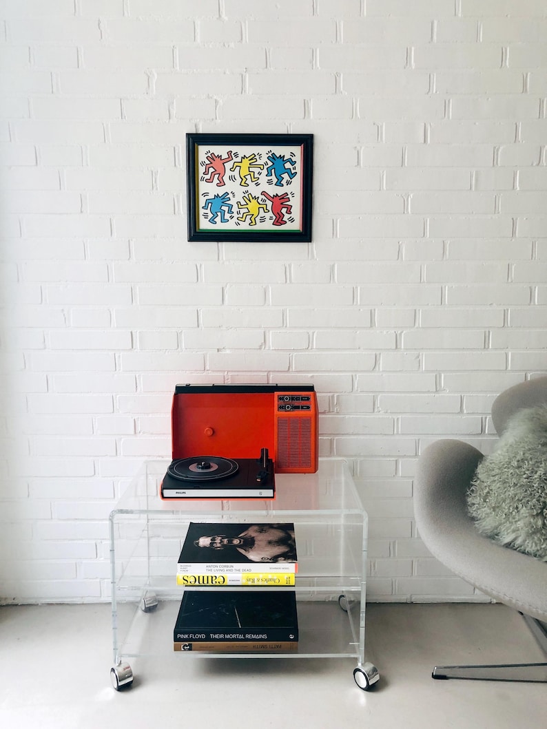 May include: A framed artwork with colorful dancing figures hangs on a white brick wall. Below, a clear acrylic rolling cart holds a vintage orange record player and a stack of books. A gray armchair with a fluffy pillow sits nearby.
