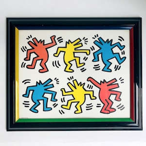 May include: Framed artwork featuring six colorful figures in red, yellow, and blue, dancing on a white background. The artwork is framed in a black frame with a red, yellow, and green inner border. The style is reminiscent of Keith Haring's work.