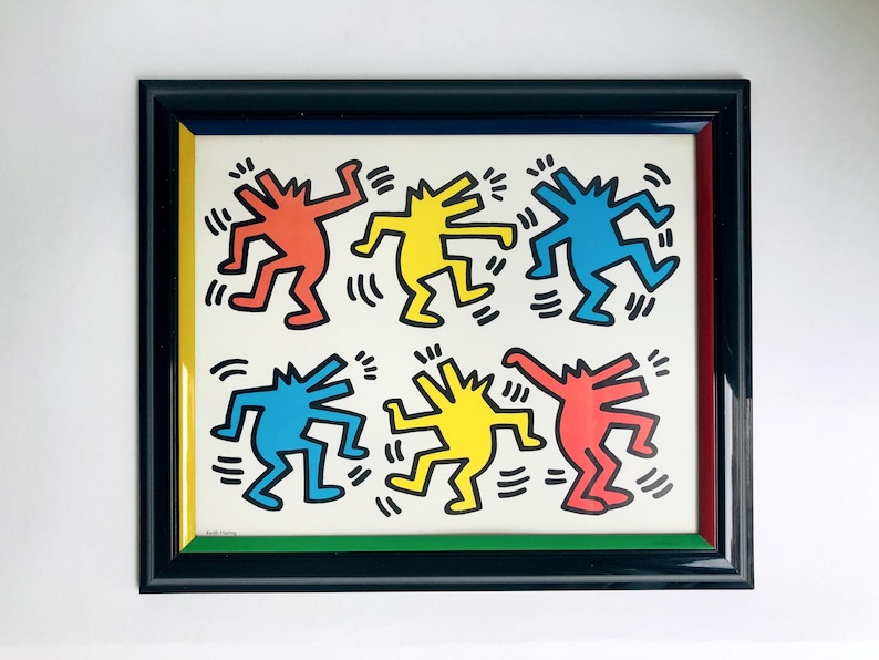 May include: Framed artwork featuring six colorful figures in red, yellow, and blue, dancing on a white background. The artwork is framed in a black frame with a red, yellow, and green inner border. The style is reminiscent of Keith Haring's work.