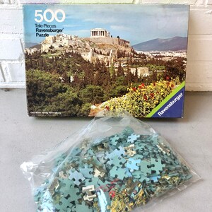 K&ouml;nnte beinhalten: Ein 500-teiliges Ravensburger Puzzle mit einem malerischen Blick auf die Akropolis in Athen, Griechenland. Die Schachtel zeigt ein farbenfrohes Bild des antiken griechischen Wahrzeichen mit &uuml;ppigen gr&uuml;nen B&auml;umen und blauem Himmel.