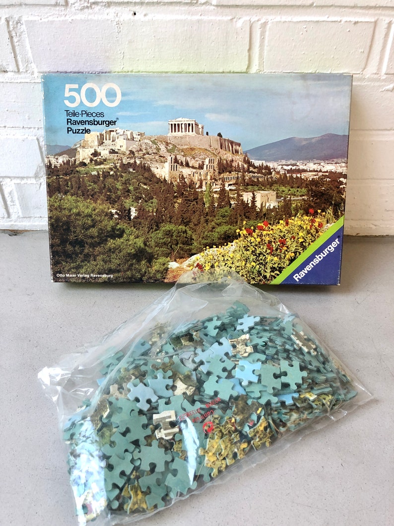 K&ouml;nnte beinhalten: Ein 500-teiliges Ravensburger Puzzle mit einem malerischen Blick auf die Akropolis in Athen, Griechenland. Die Schachtel zeigt ein farbenfrohes Bild des antiken griechischen Wahrzeichen mit &uuml;ppigen gr&uuml;nen B&auml;umen und blauem Himmel.