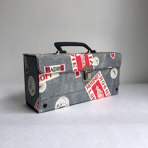 May include: A denim-effect box with a black handle and a silver clasp. The box is adorned with red and white travel-themed stickers, including the words "Madrid" and "London". The box is designed for carrying items.