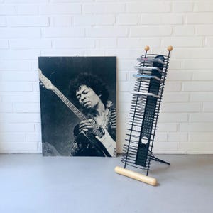 May include: A black and white poster of Jimi Hendrix playing a guitar is next to a black metal CD rack. The CD rack has a wooden base and top accents. The rack is holding several CDs.