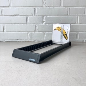 May include: A black CD rack with a rectangular frame and multiple vertical dividers. A CD case with a yellow banana graphic and the text "MY GENERATION" is displayed in the rack. The brand name "hama" is visible on the front.