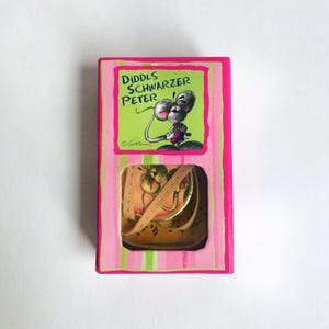 Könnte beinhalten: Eine rechteckige Schachtel mit rosa und grün gestreiftem Rand, mit dem Text "Diddls Schwarzer Peter" und einer Cartoon-Maus-Illustration. Die Schachtel enthält ein Vintage-Maßband.