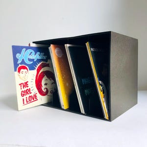 以下が含まれることがあります： 黒いレコード収納ボックスには、いくつかのアルバムカバーが入っています。 アルバムカバーには、「THE GIRL I LOVE」や「MASTER MAG」などのテキストを含む、さまざまなデザインと色があります。 ボックスは、暗く、質感のある素材で作られています。