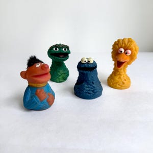 Könnte beinhalten: Ein Set aus vier Sesame Street Fingerpuppen aus geformtem Gummi. Die Sammlung umfasst eine orangefarbene Figur mit schwarzen Haaren, eine grüne texturierte Figur, eine blaue, fellartig strukturierte Figur mit großen Augen und eine gelbe Vogel-Figur.
