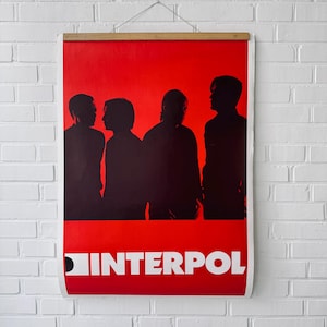 May include: A red poster with the band Interpol in white text at the bottom. The poster features black silhouettes of four people against a red background. The poster is held by a wooden frame.