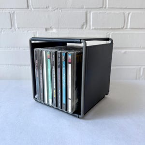 May include: A black CD storage rack holding several CDs. The rack has a square shape with a metal frame and a solid side. The CDs are visible, with their cases and album art displayed. The rack is on a white surface.