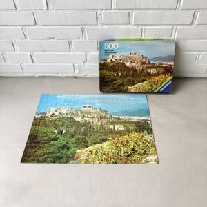 K&ouml;nnte beinhalten: Ein 500-teiliges Ravensburger Puzzle mit einem malerischen Blick auf die Akropolis in Athen, Griechenland. Die Schachtel des Puzzles zeigt ein farbenfrohes Bild der Akropolis mit &uuml;ppigen gr&uuml;nen B&auml;umen und blauem Himmel.