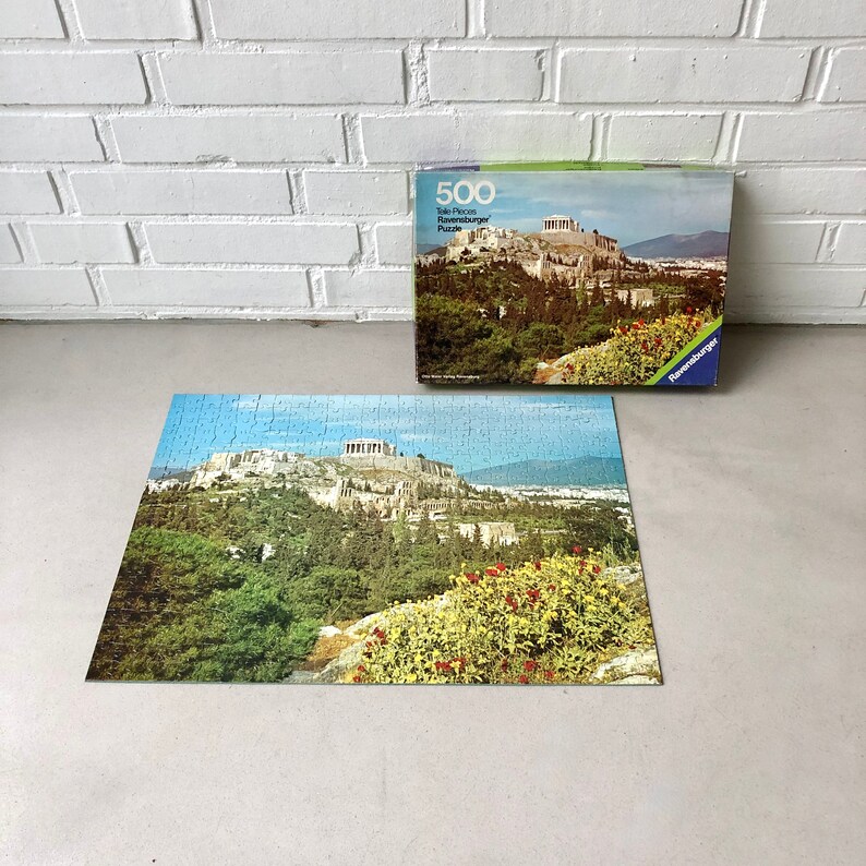 K&ouml;nnte beinhalten: Ein 500-teiliges Ravensburger Puzzle mit einem malerischen Blick auf die Akropolis in Athen, Griechenland. Die Schachtel des Puzzles zeigt ein farbenfrohes Bild der Akropolis mit &uuml;ppigen gr&uuml;nen B&auml;umen und blauem Himmel.