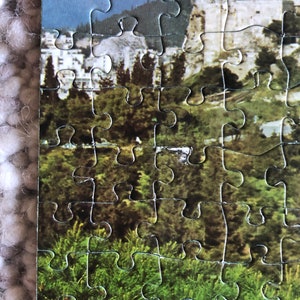 K&ouml;nnte beinhalten: Eine Nahaufnahme eines teilweise zusammengesetzten Puzzles, das eine Landschaft mit einer Bergkette, einer Stadt und einem Wald im Vordergrund zeigt.