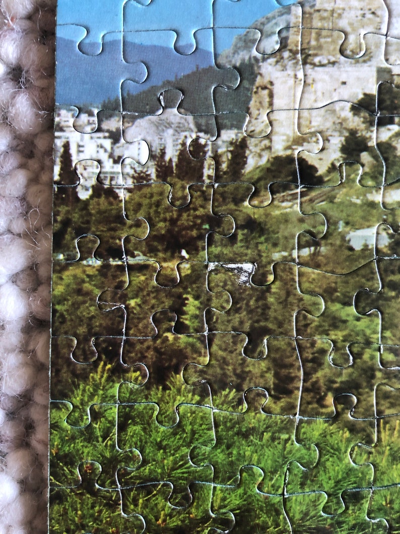 K&ouml;nnte beinhalten: Eine Nahaufnahme eines teilweise zusammengesetzten Puzzles, das eine Landschaft mit einer Bergkette, einer Stadt und einem Wald im Vordergrund zeigt.