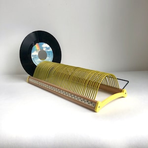 Könnte beinhalten: Ein gelber Schallplattenständer aus Draht mit einem Holzsockel, der eine aufgedruckte Zahlenskala von 1 bis 50 aufweist. Eine einzelne schwarze 7-Zoll-Schallplatte mit einem Regenbogen-Label befindet sich im ersten Schlitz des Metallhalters.